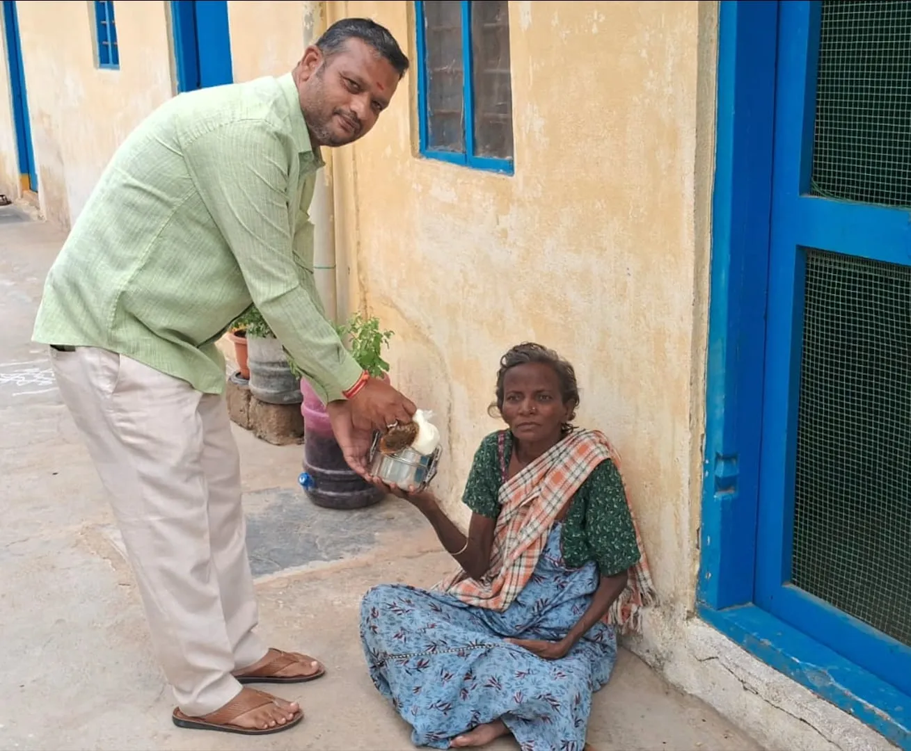 A volunteer kneeling to hand a freshly cooked meal to an elderly woman outside her home in Puttaparthi